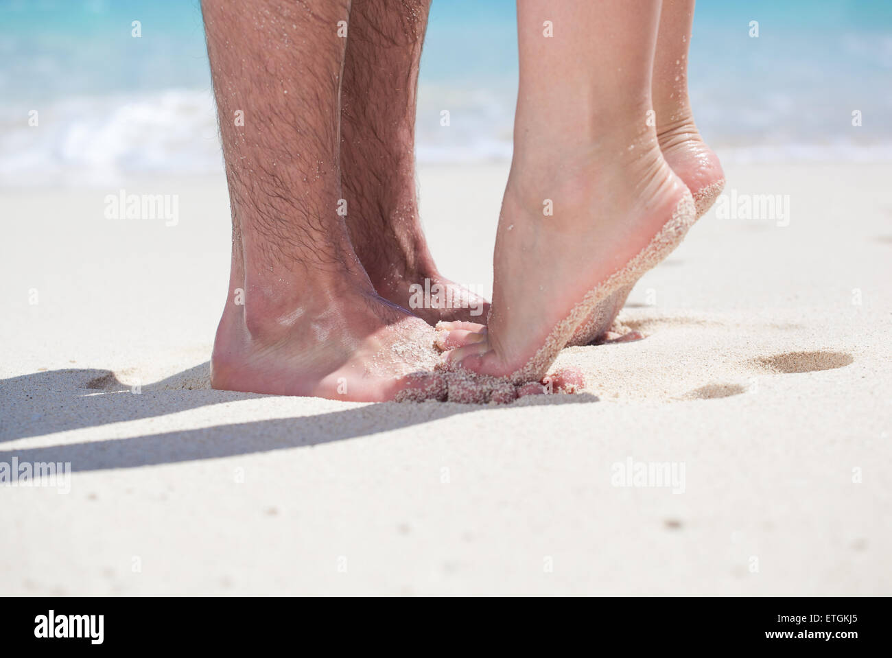 Woman's barefoot legs standing up tiptoe closeup to male foot and kissing on sandy beach with turquoise sea background, no face. Stock Photo
