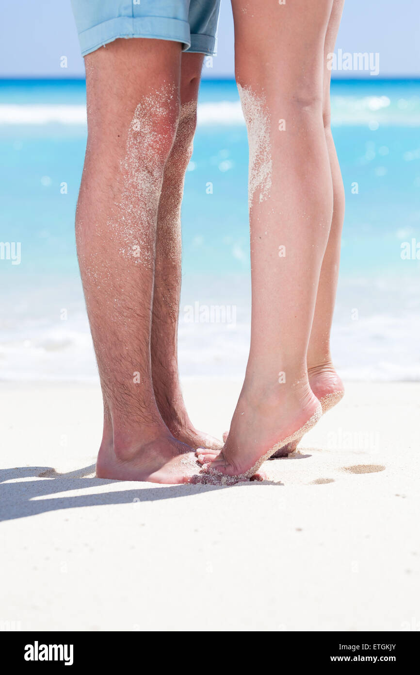 Woman's barefoot legs standing up tiptoe closeup to male foot and kissing on sandy beach with turquoise sea background, no face. Stock Photo