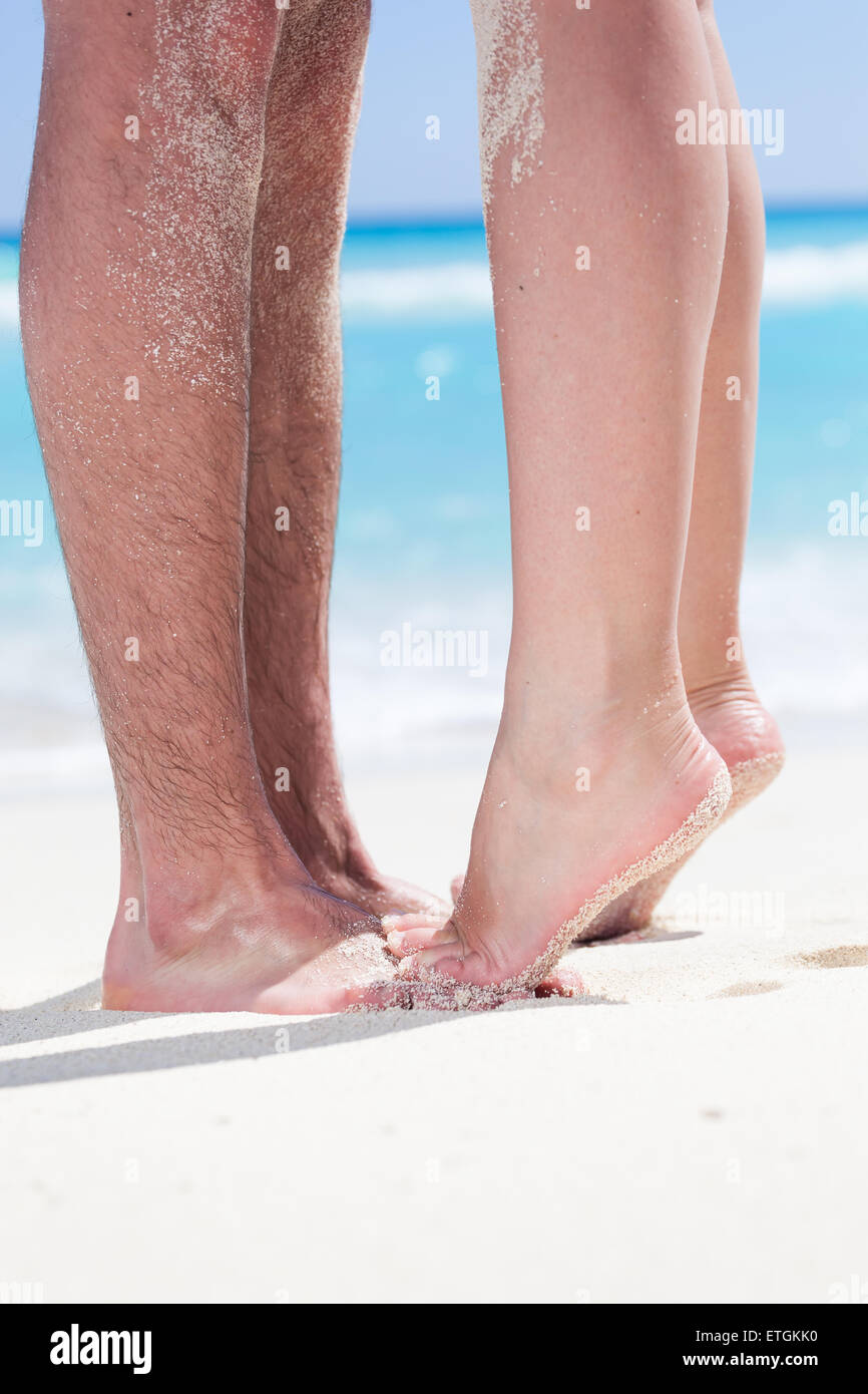 Woman's barefoot legs standing up tiptoe closeup to male foot and kissing on sandy beach with turquoise sea background, no face. Stock Photo