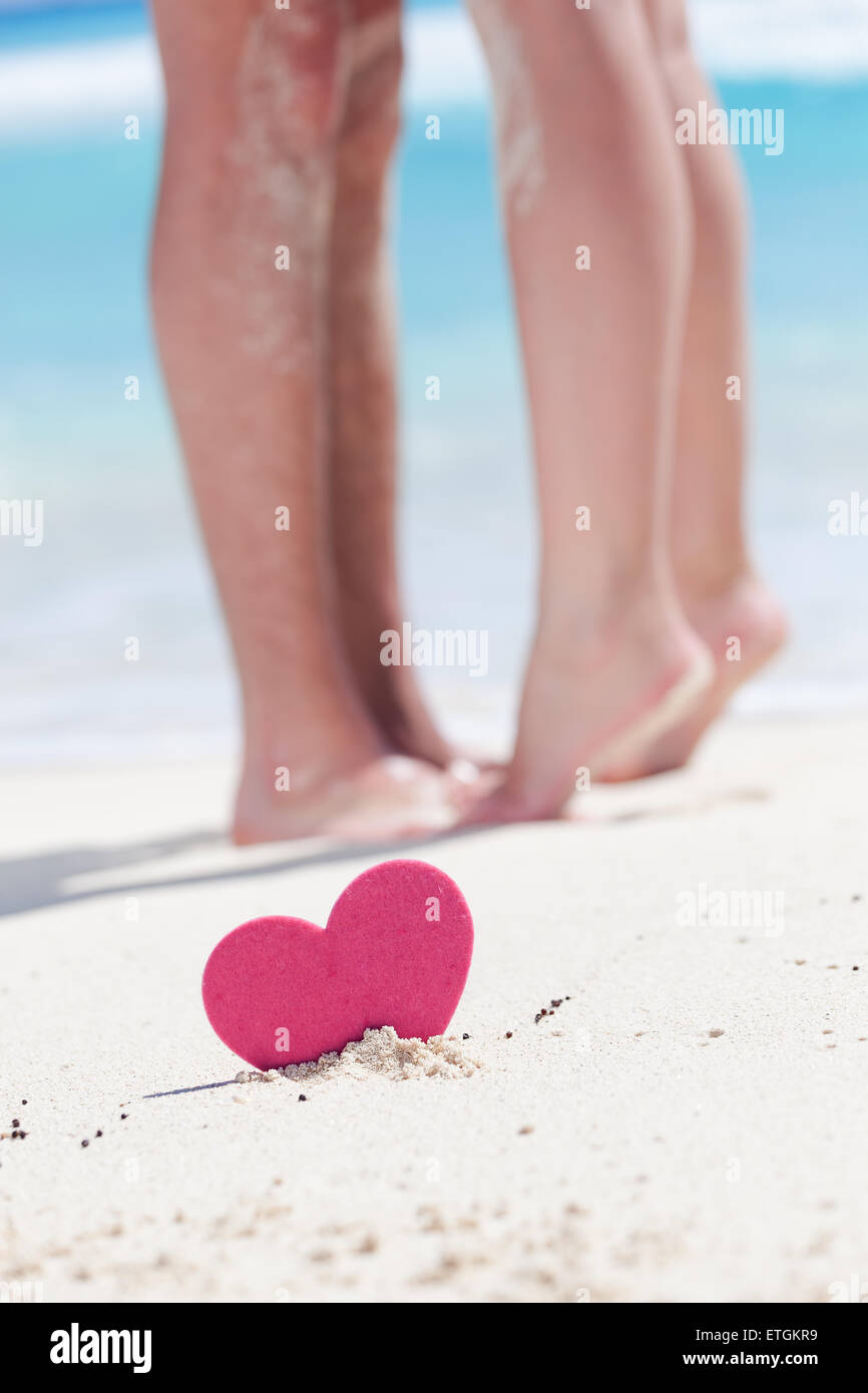Barefoot female legs standing up tiptoe on man's foots on beach with turquoise sea background, decorated pink heart object.  Rom Stock Photo