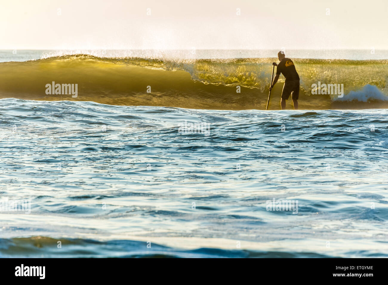 A stand-up paddleboarder faces a backlit wave as he paddle out at Jacksonville Beach, Florida just after sunrise. Stock Photo