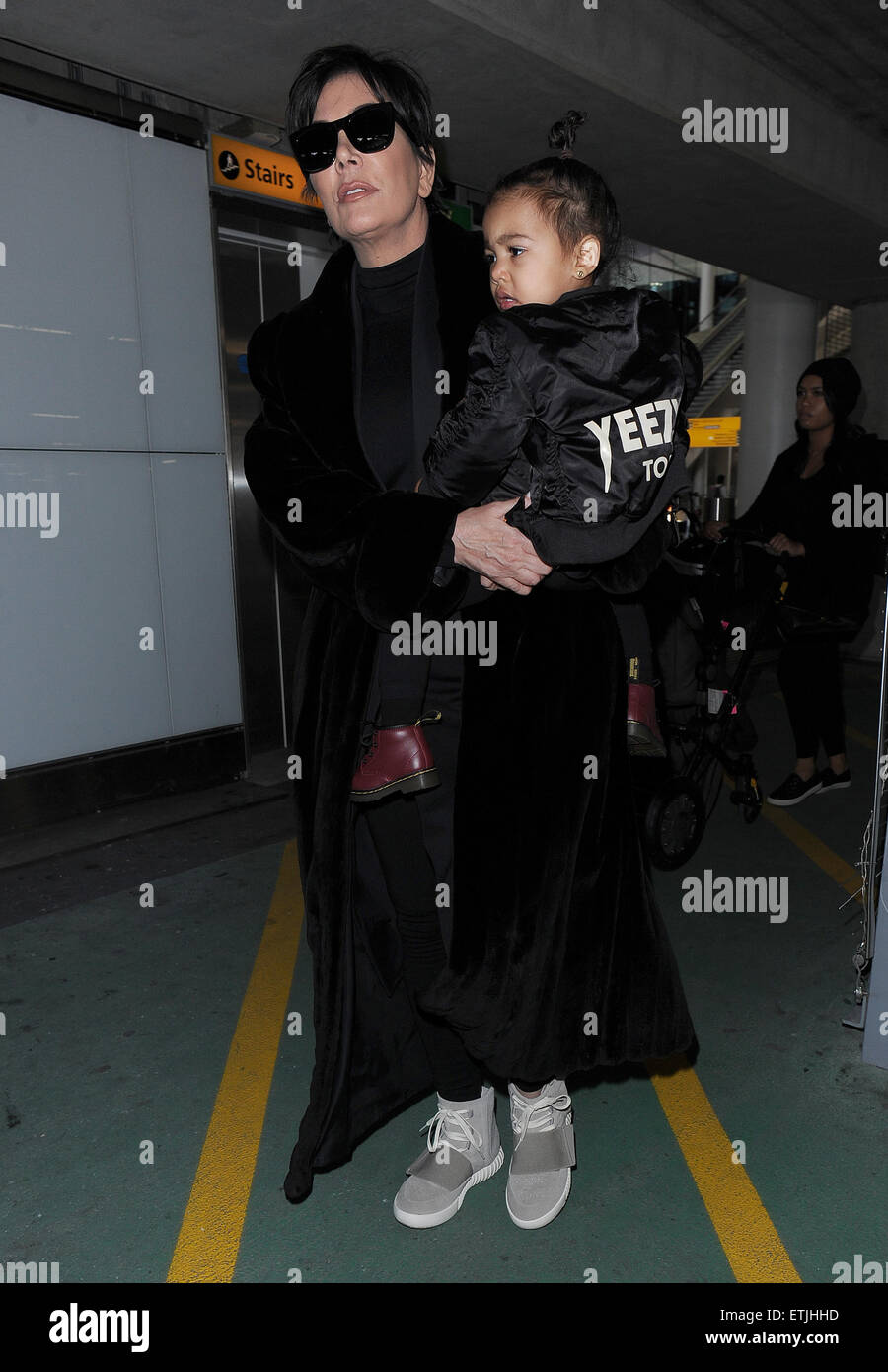 Kris Jenner wearing a pair of Kanye West x Adidas Originals Yeezy 750 Boost arrives at Heathrow Airport carrying baby North West who was wearing a Yeezus Tour bomber jacket in support