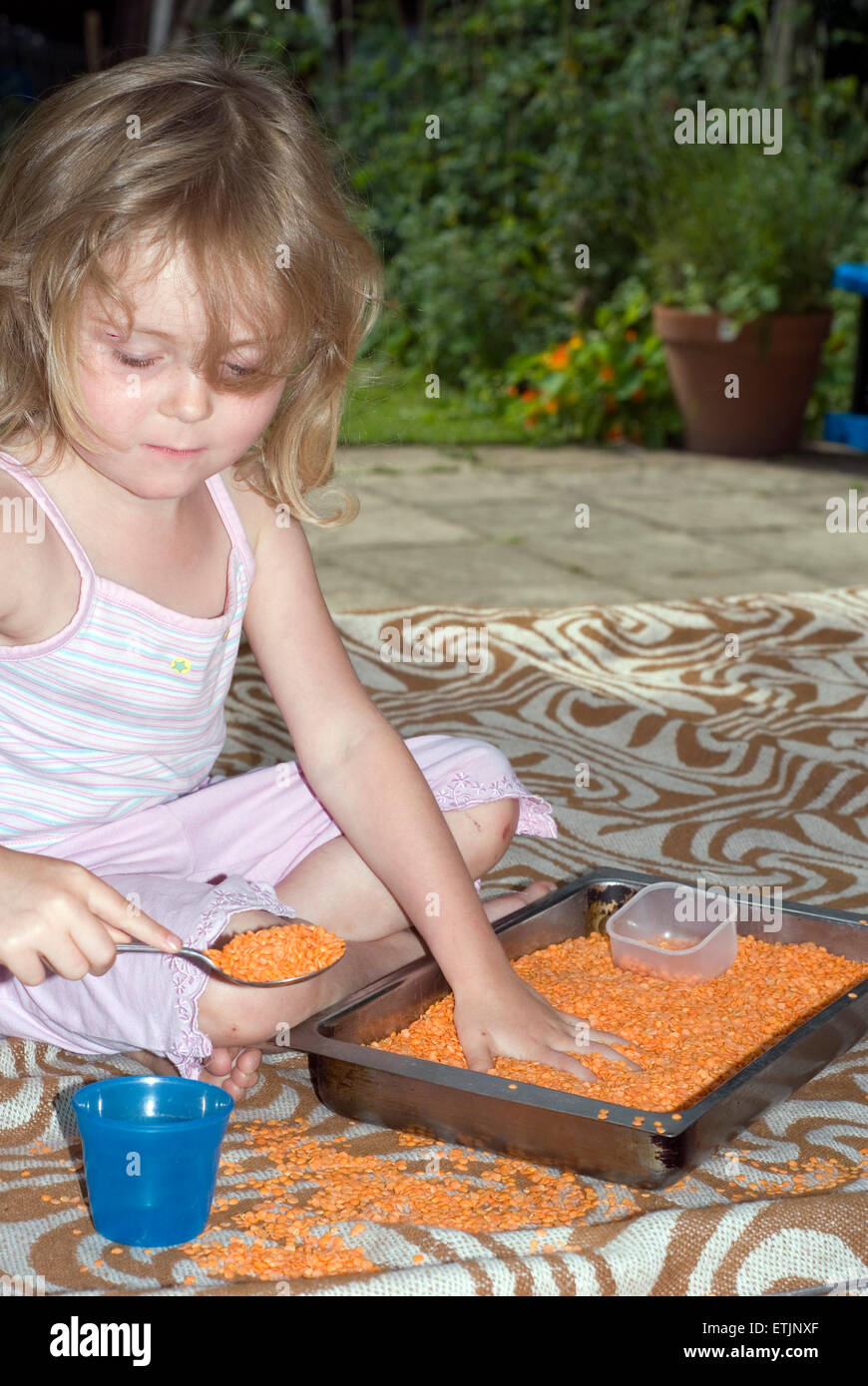 Little girl playing tactile games with a tray of lentils and some pots and spoons outdoors in the shade on a summer day, UK Stock Photo