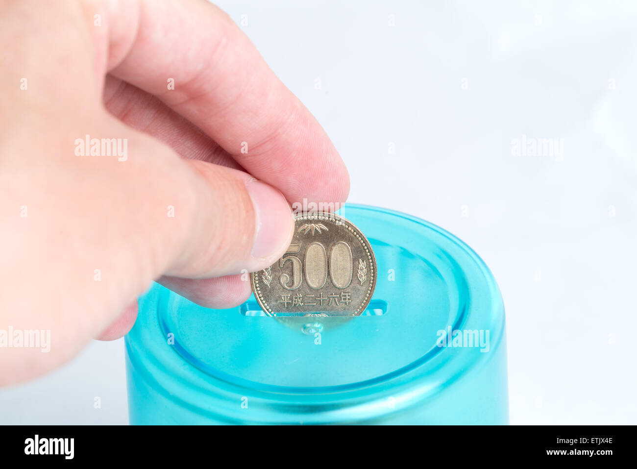 A man's hand putting a 500 Japanese Yen coin into a blue bank with a white background. Stock Photo