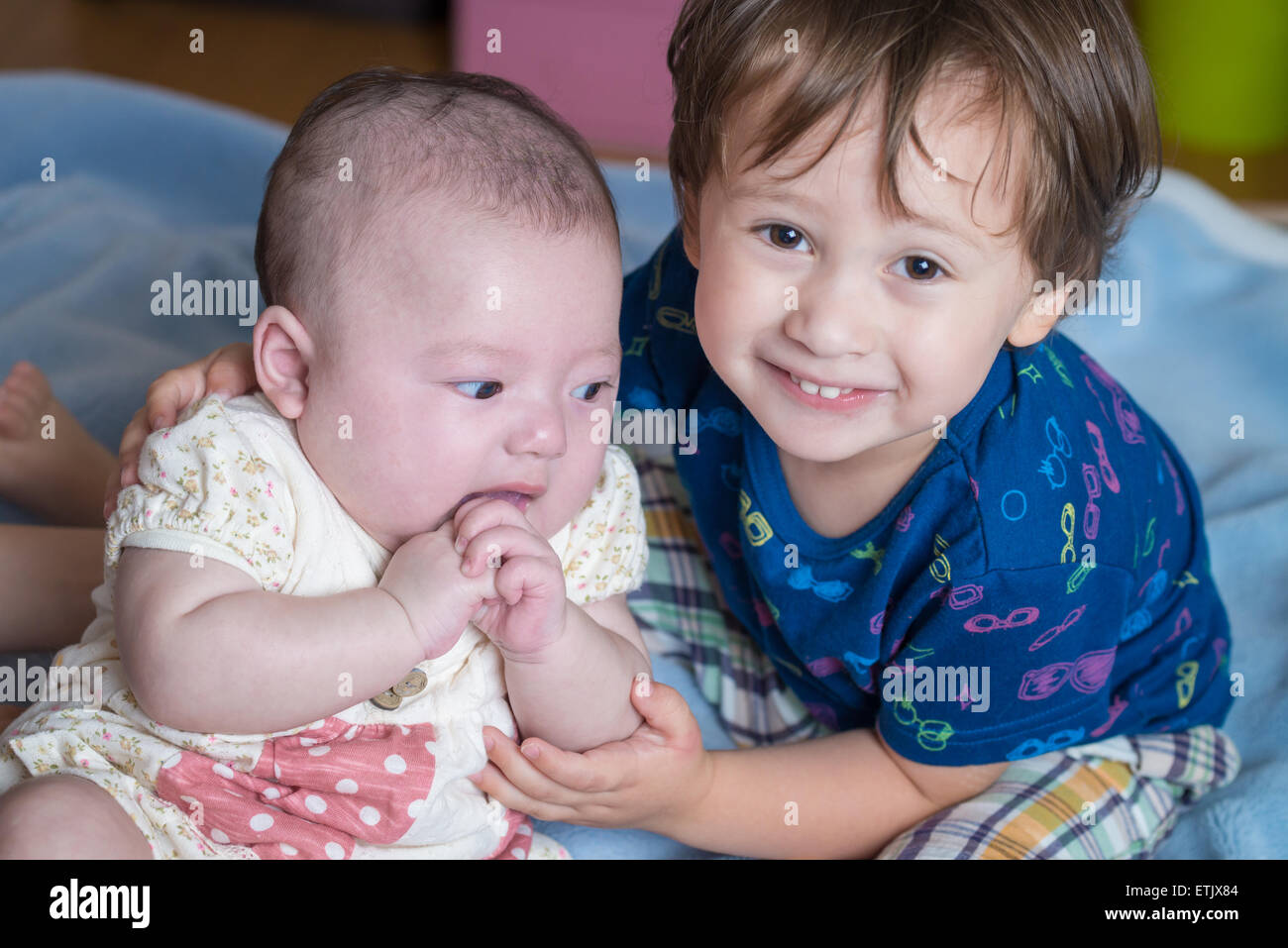 A 2 year old boy and his newborn sister sitting on a bed. Stock Photo