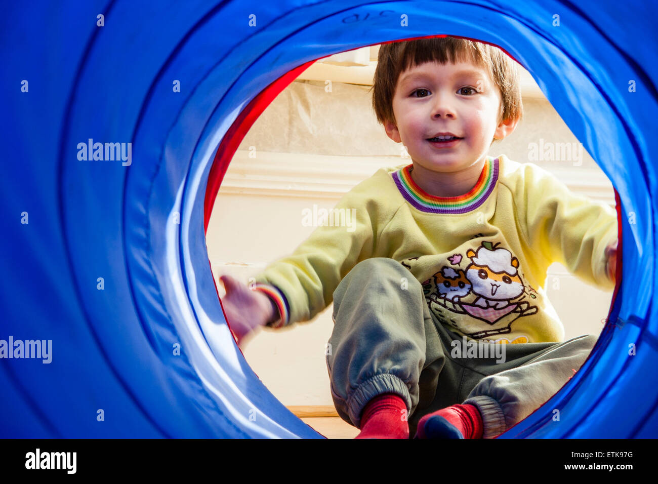 View from inside blue fabric play tunnel of young child, boy, 4-5 year old, at end of the tunnel, holding edges and looking at viewer, smiling. Stock Photo