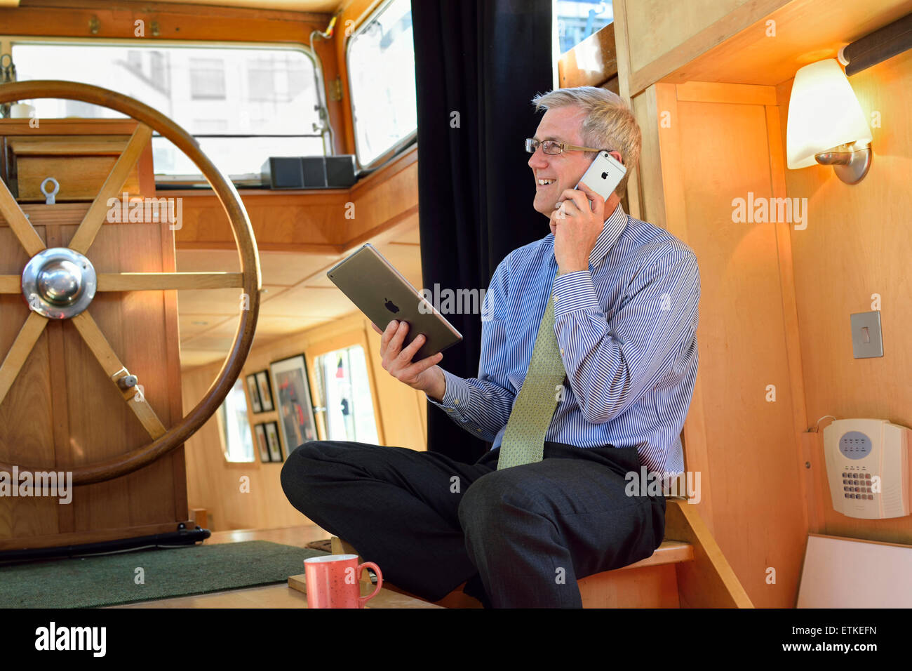 Relaxed mature businessman in houseboat office holding his iPad air tablet computer screen and using his iPhone 6 smartphone Stock Photo