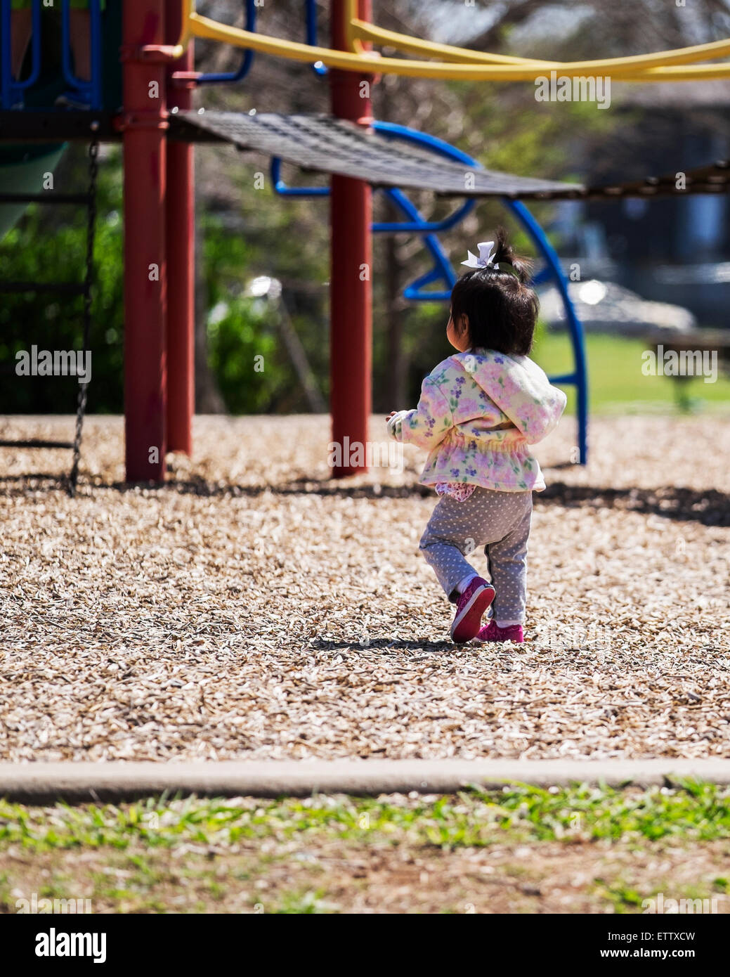 A one year old ethnic baby girl toddles toward playground equipment. USA. Stock Photo