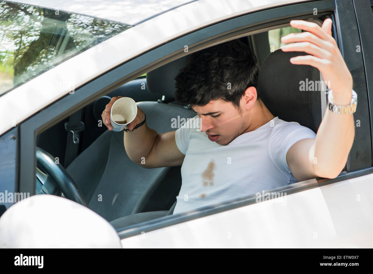 Young Man Having a Bad Day, Distracted Driver Looking Down in Frustration at Spilled Coffee on White T-Shirt While Sitting in Dr Stock Photo