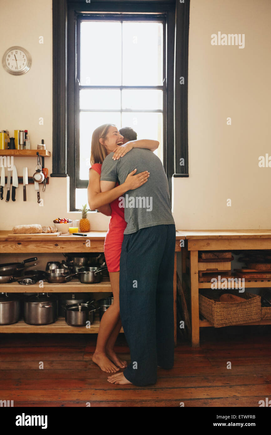 Happy young woman being hugged by her boyfriend in the kitchen. Cheerful young couple embracing each other in morning at home. Stock Photo