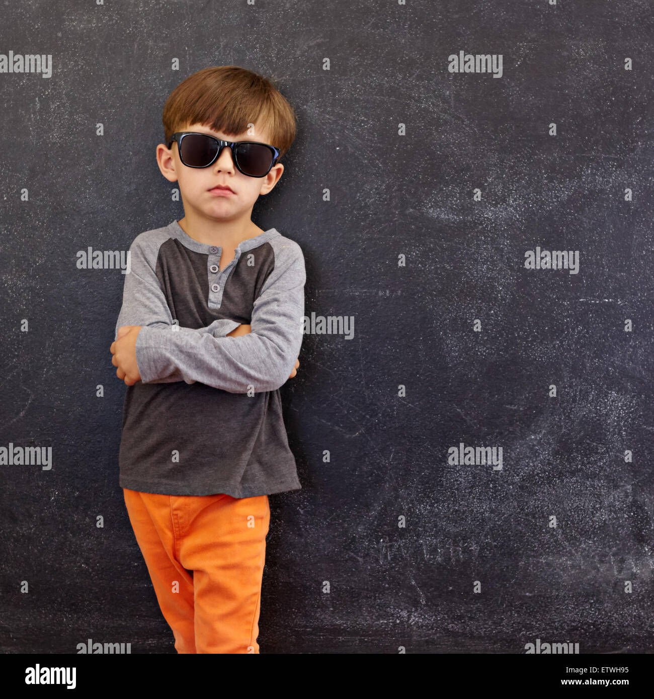 Portrait of smart little boy wearing sunglasses standing with his hands folded against blackboard with copyspace. Stock Photo