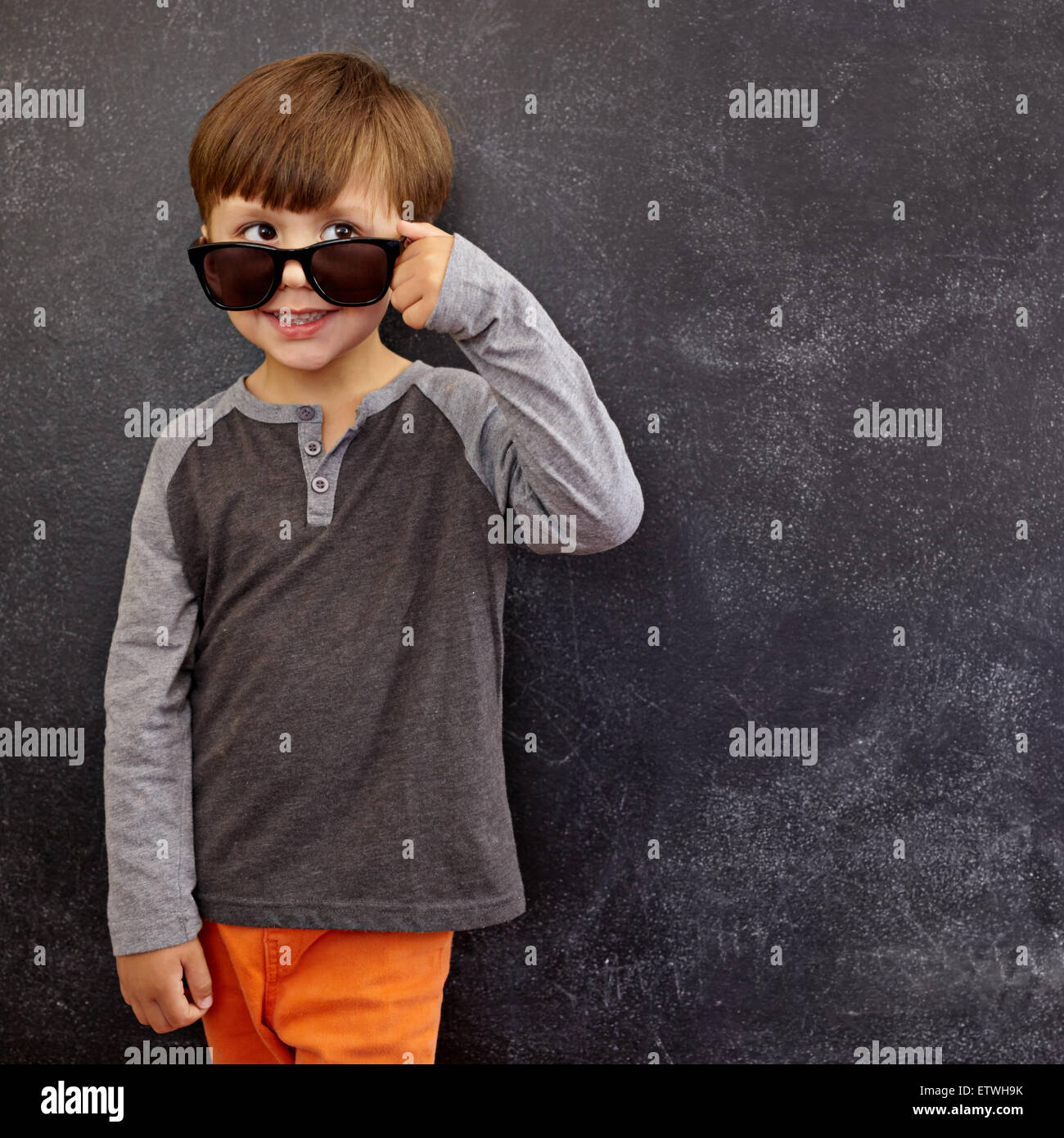 Portrait of a little boy in sunglasses looking at away at copy space. Cool boy peering over his sunglasses against blackboard. Stock Photo