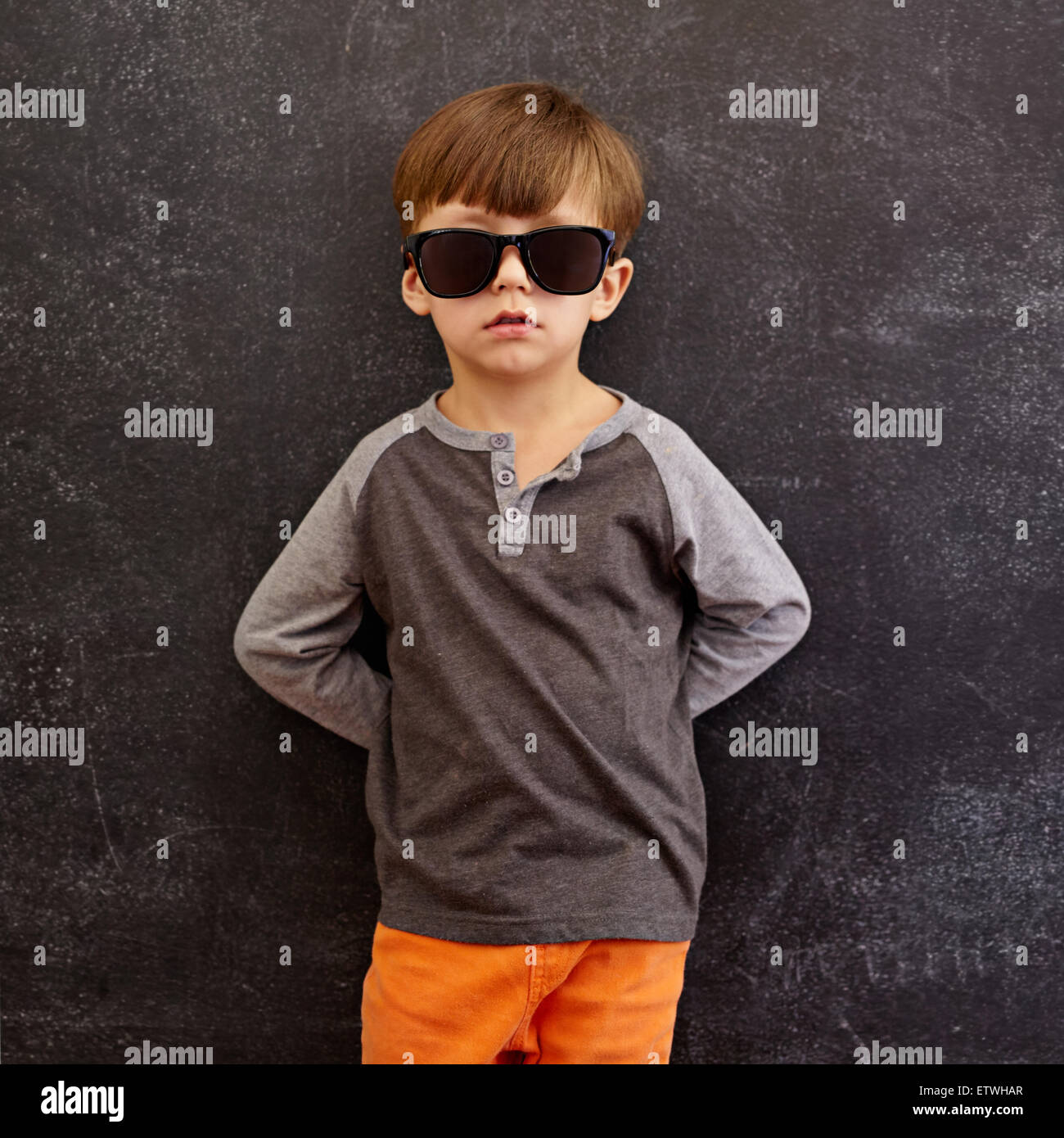 Portrait of innocent little kid wearing sunglasses. Little boy leaning on a blackboard. Square composition. Stock Photo