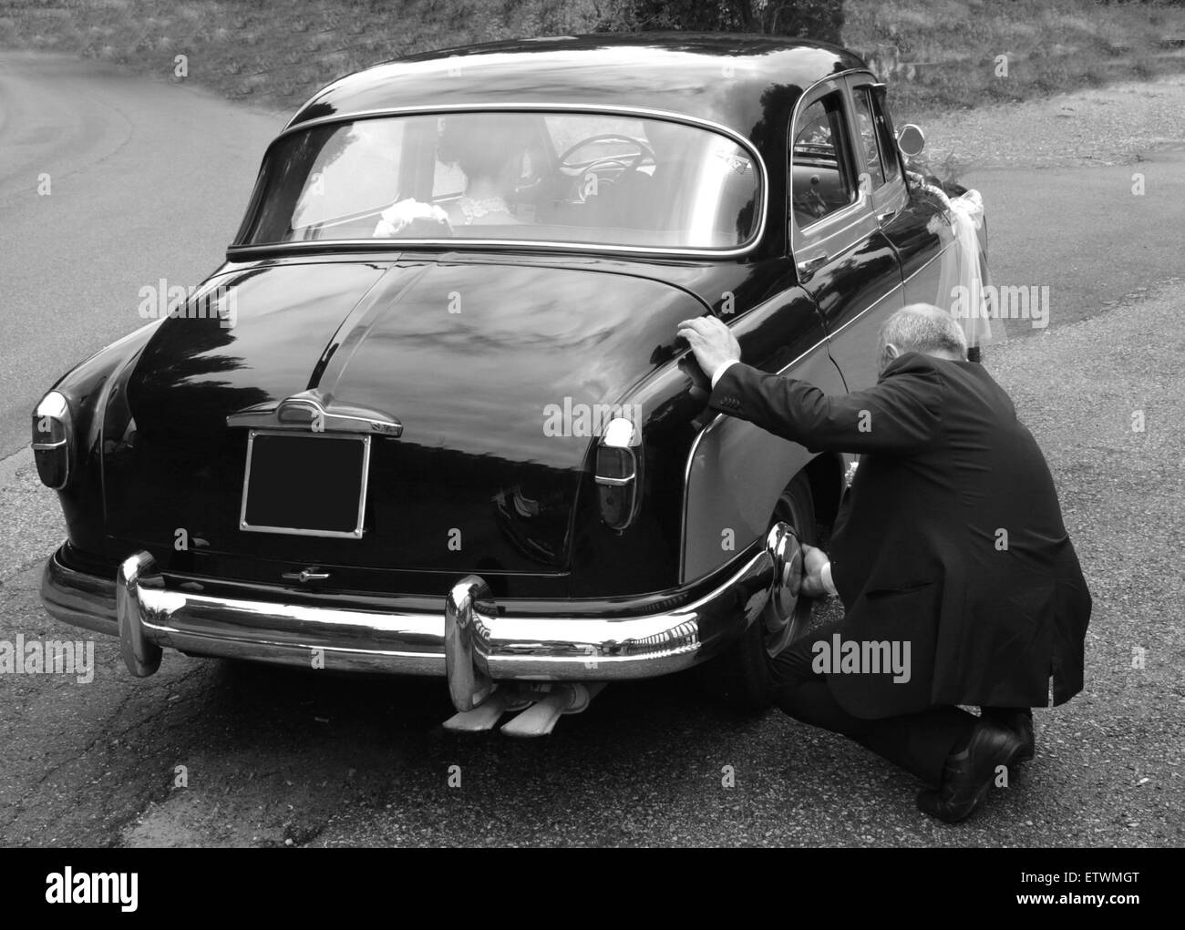 an old driver repairing retro wedding car while bride and groom kissing inside Stock Photo