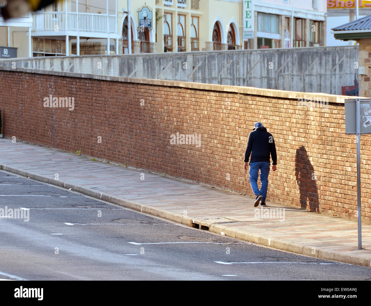 Man walking along a pavement next to a brick wall in a patch of sunlight Stock Photo
