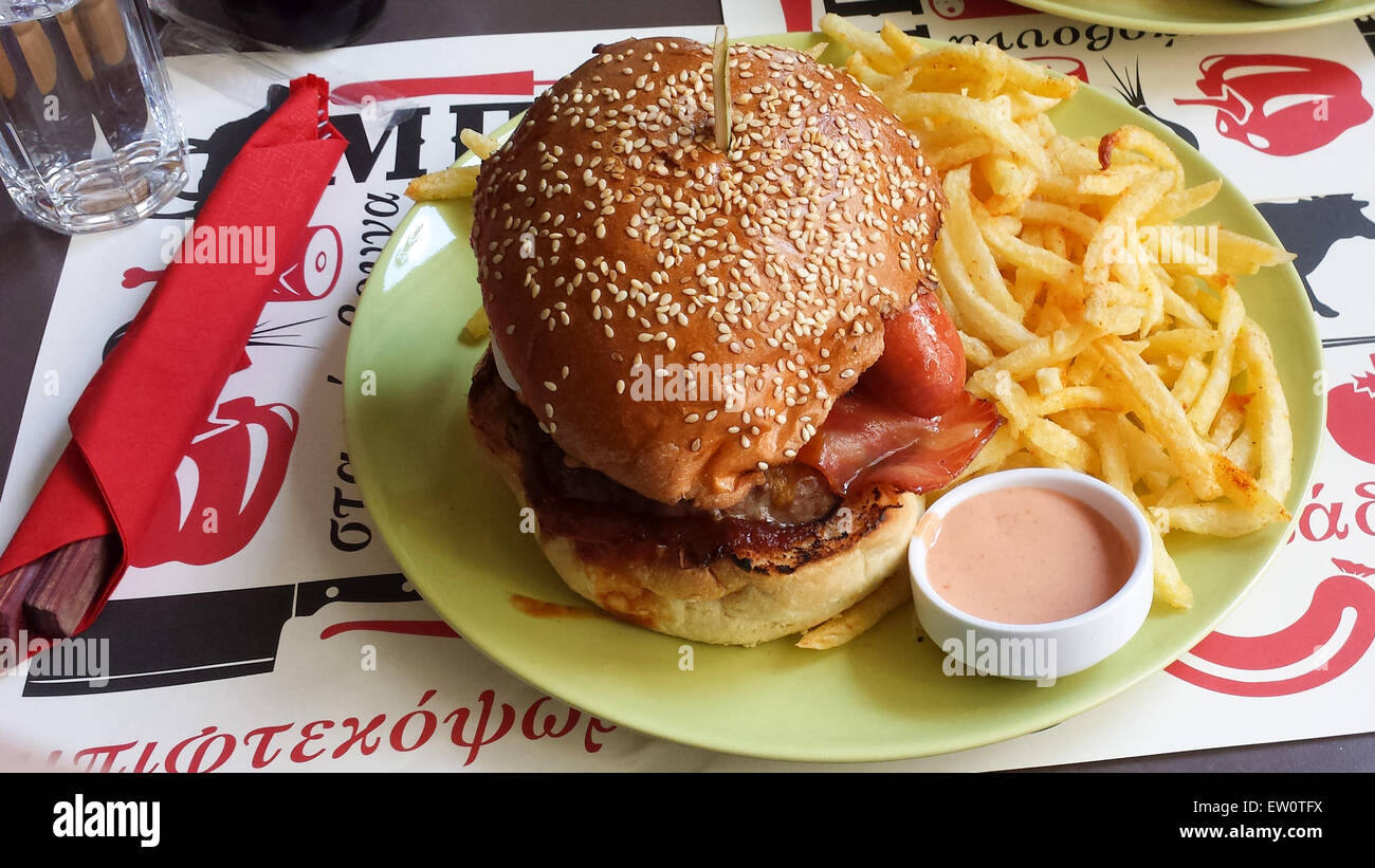 View of a huge hamburger with burger, fried eggs, sausage, barbecue sauce and bacon accompanied by fried potatoes and sauce. Stock Photo