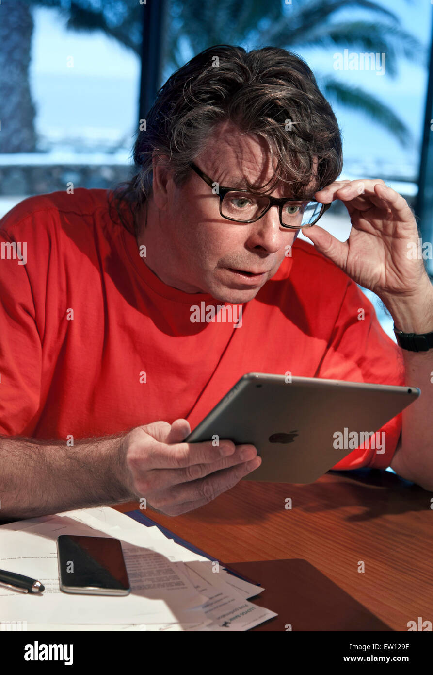 Man in holiday setting surprised by detail screen information on his iPad Air tablet computer with iPhone 6 smartphone nearby Stock Photo
