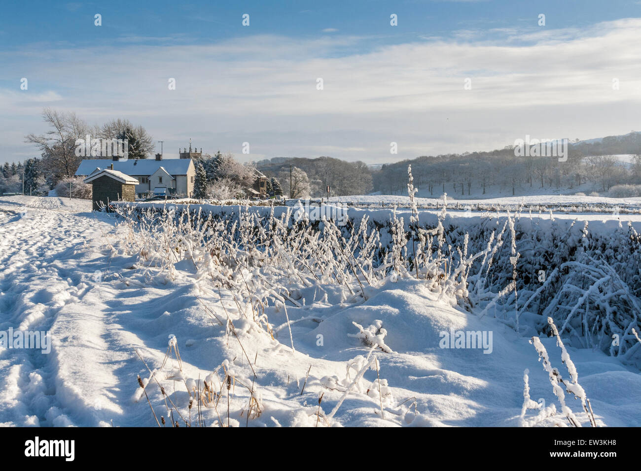 Drifts of snow in the countryside leading into a snow covered house in the distance with pale blue skies Stock Photo