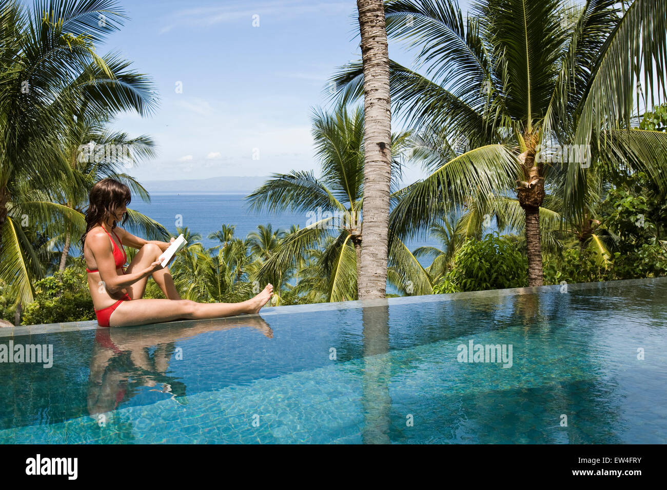 A woman relaxing by the pool reading a book under a palm tree. Stock Photo