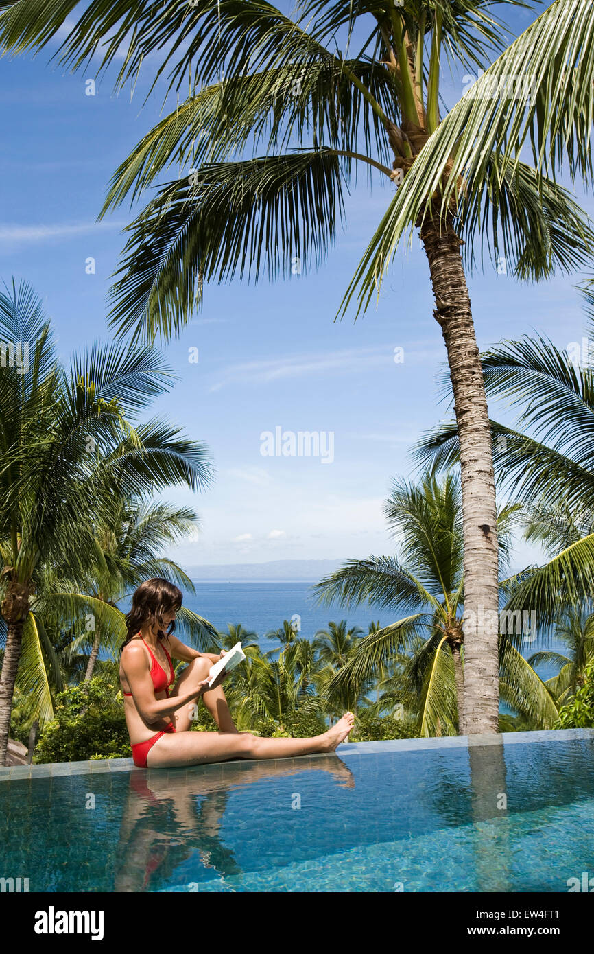 A woman relaxing by the pool reading a book under a palm tree. Stock Photo