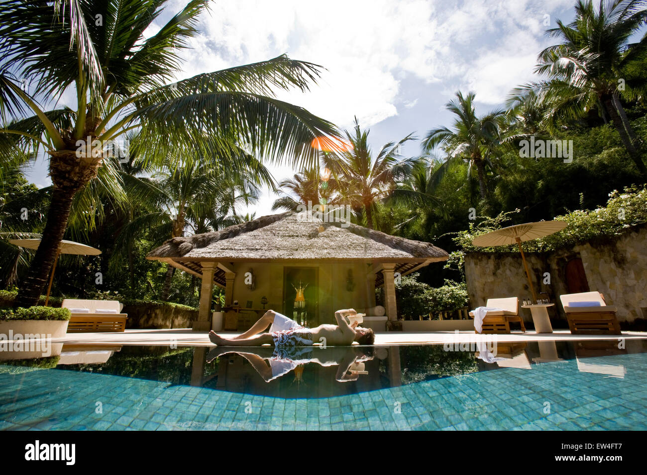 A man relaxing by the pool of his bungalow reading a book under a palm tree. Stock Photo
