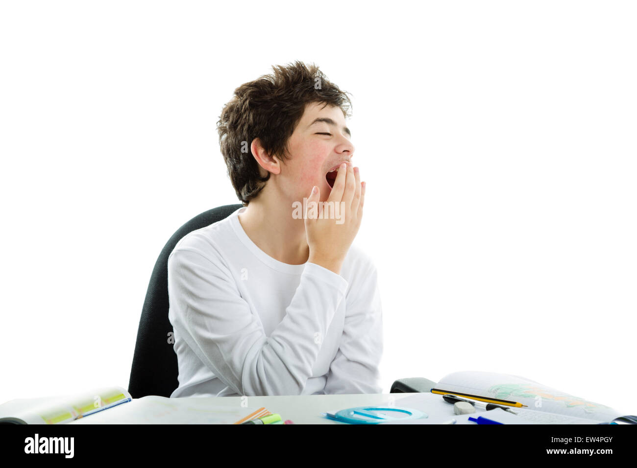 Tired Caucasian boy sits in front of homework wearing a white long sleeve t-shirt and yawns covering his mouth with right hand Stock Photo