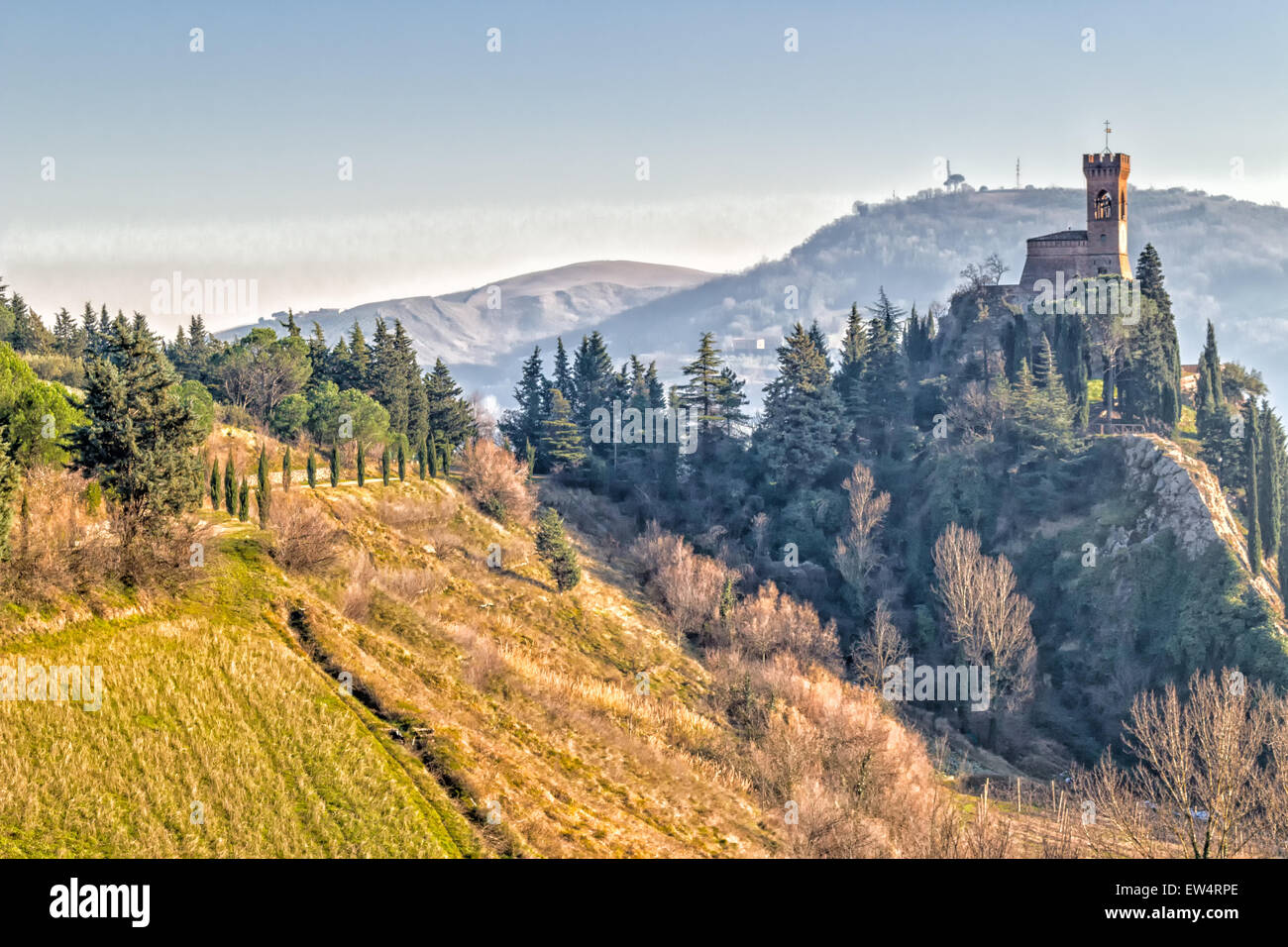 A road with cypresses trees leads to a medieval clock tower on misty hills in a countryside of yellow and reddish bushes and green cypresses in a winter sunny day in Italy Stock Photo