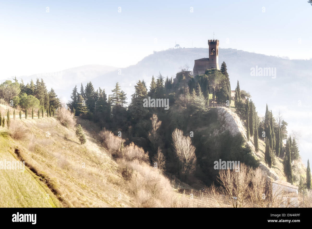 A road with cypresses trees leads to a medieval clock tower on misty hills in a countryside of yellow and reddish bushes and green cypresses in a winter sunny day in Italy Stock Photo