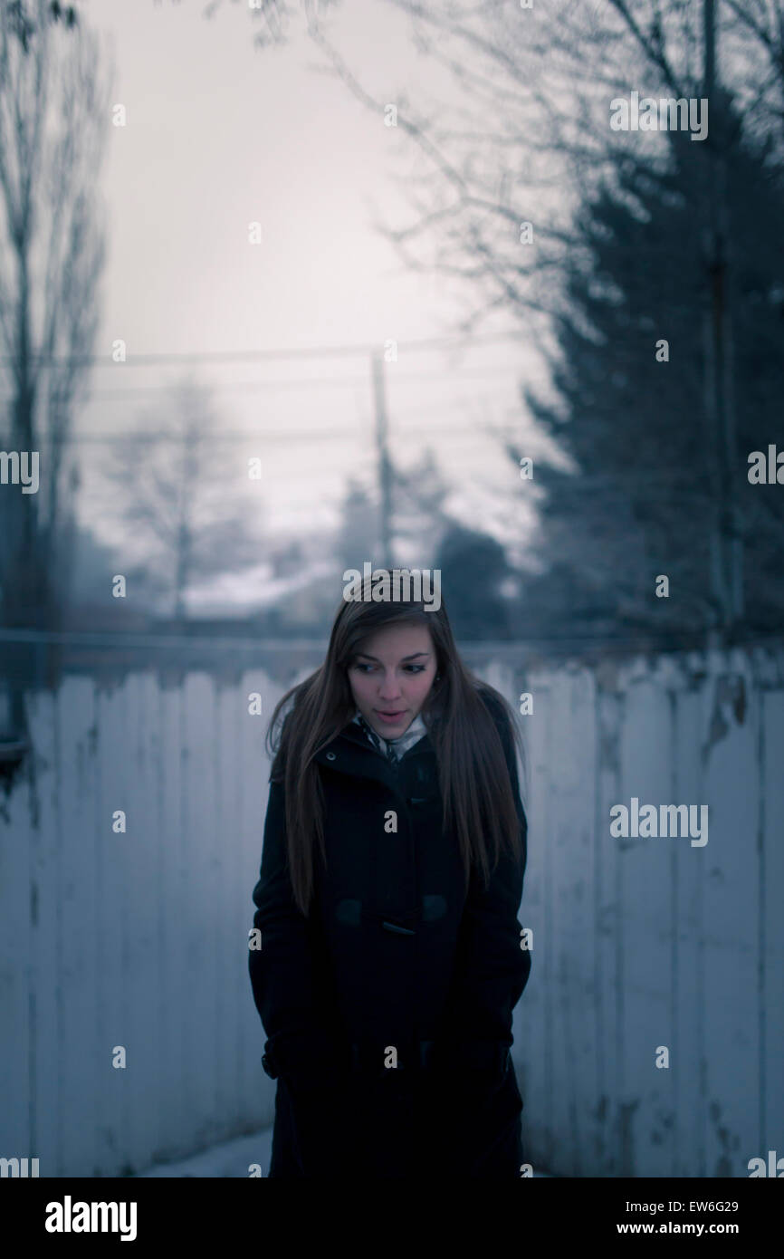 A young woman stands in front of a paint chipped fence on a foggy, dark morning. Stock Photo