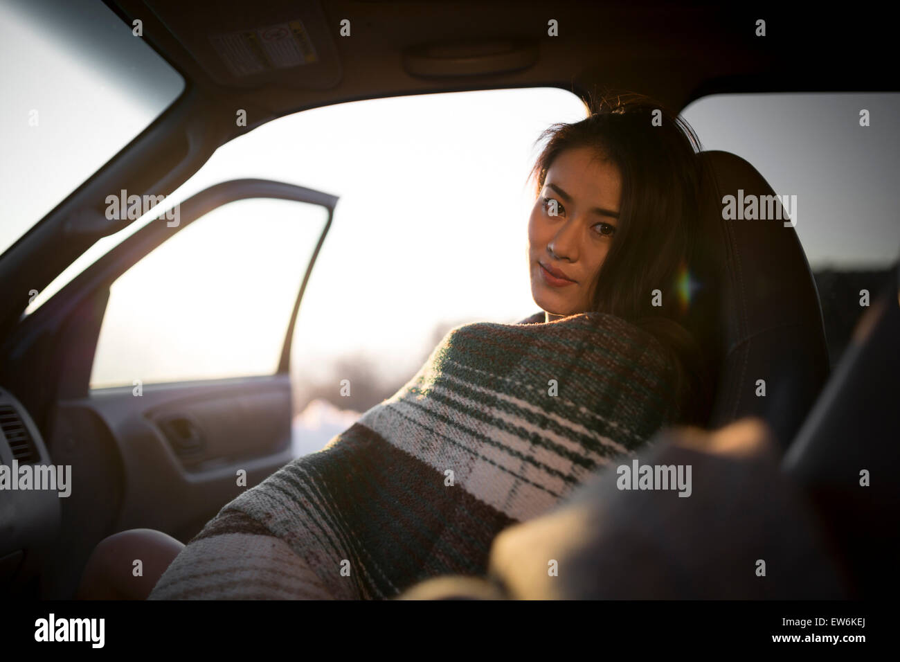 An Asian girl smiles wrapped in a blanket in a car at sunset. Stock Photo