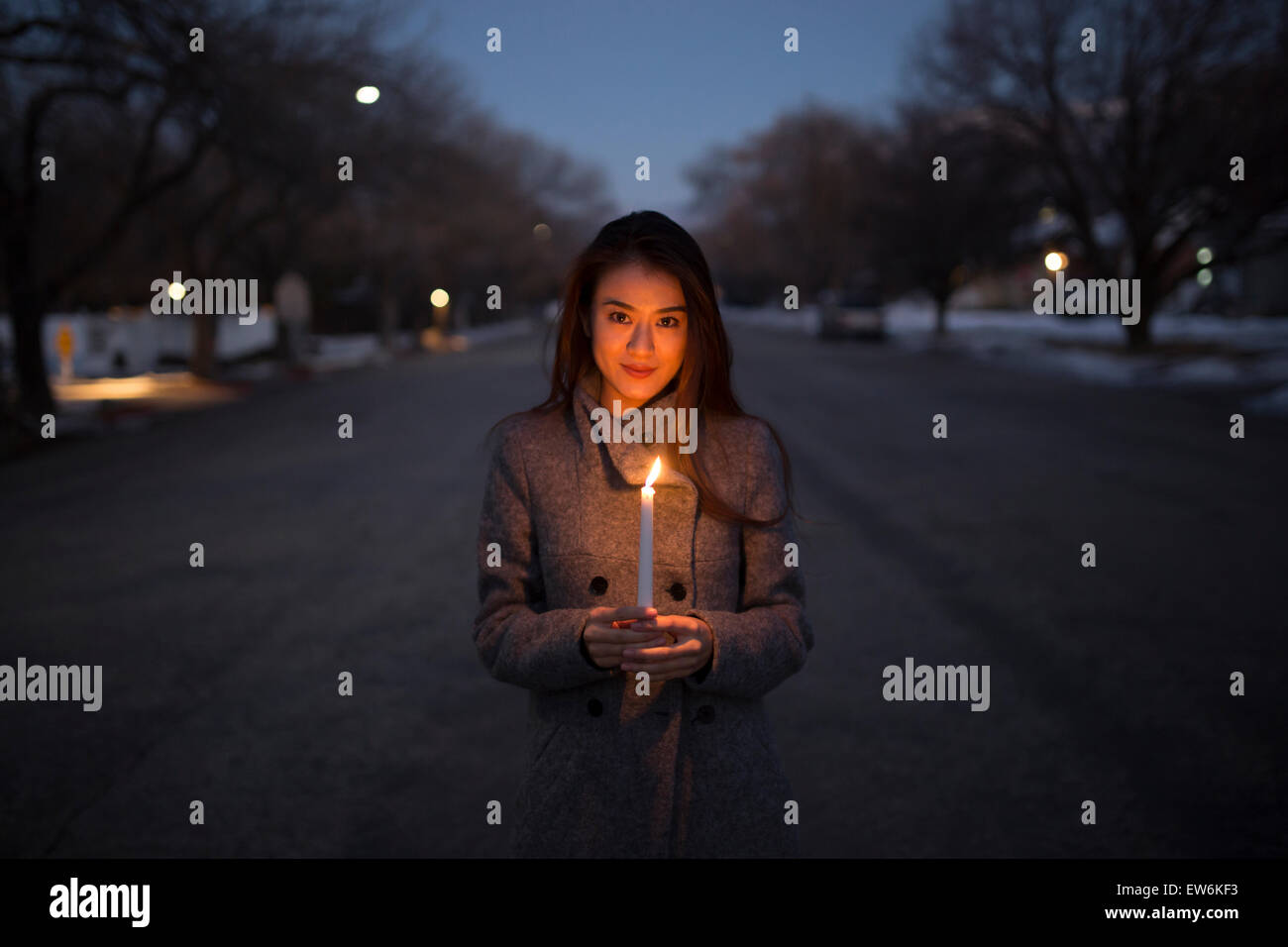An Asian girl in a coat holds a candle at dusk in the road of a small town environment. Stock Photo