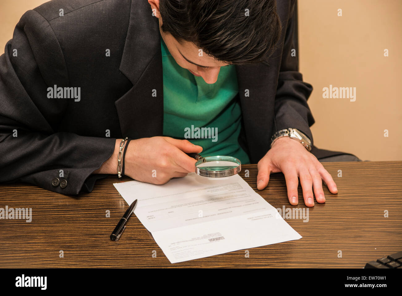 Young Man Sitting at Desk Scrutinizing Paper Contract with Magnifying Glass Prior to Signing Stock Photo