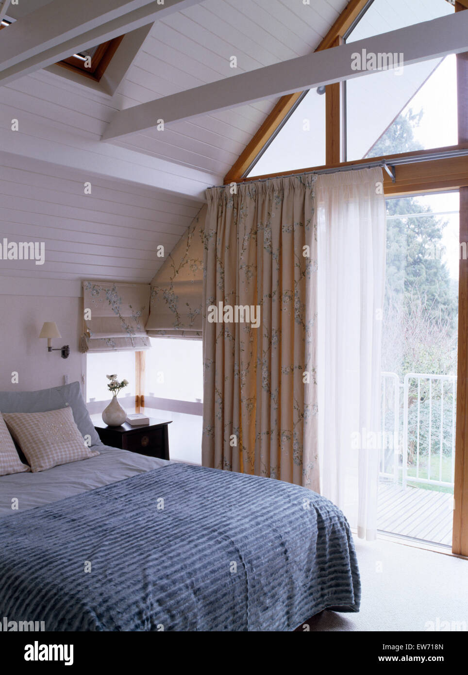 Blue-gray quilt on bed in modern loft conversion bedroom with floral drapes and white voile curtains at glass doors to balcony Stock Photo