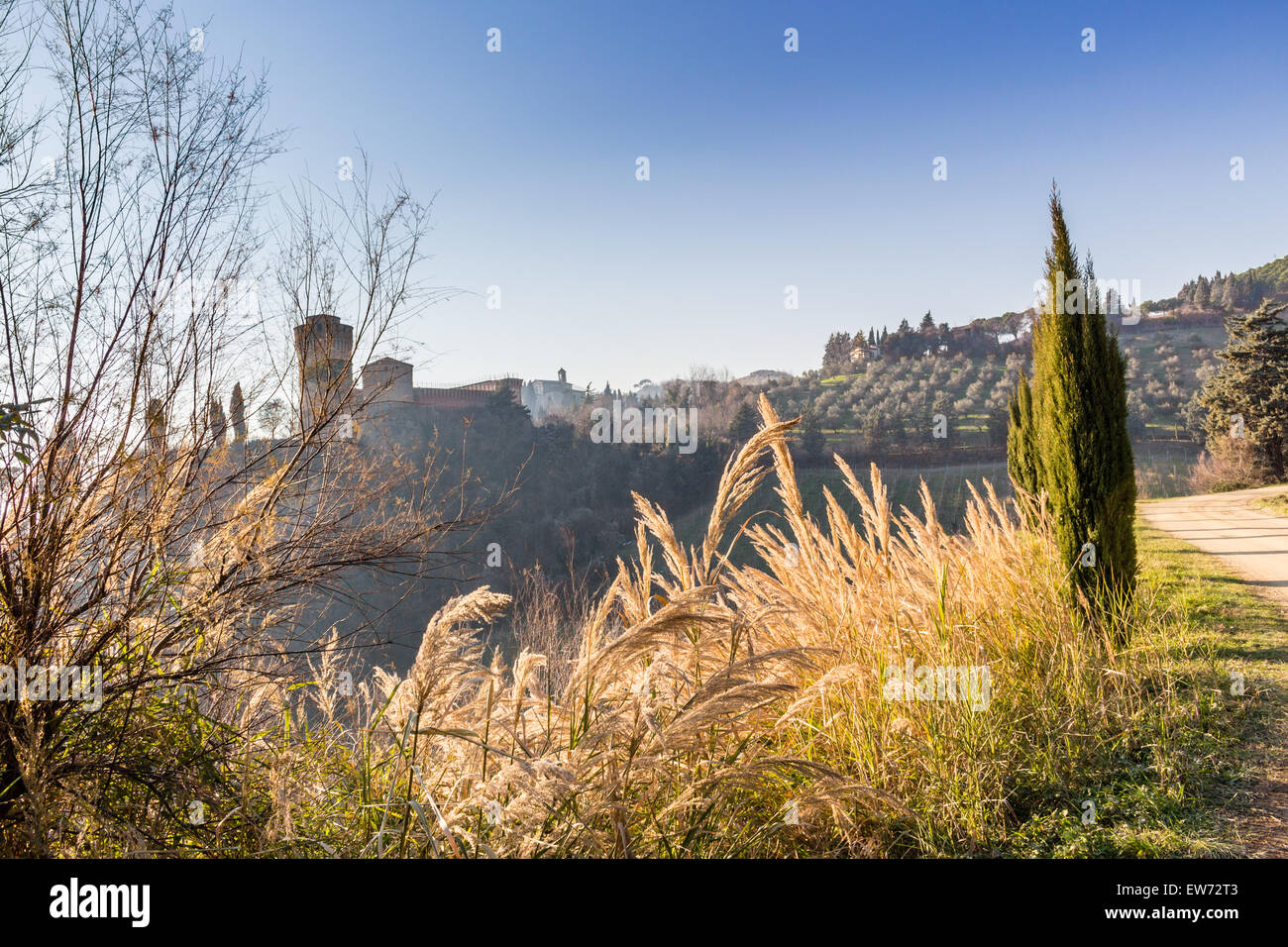 The brick walls of a medieval fortress and a sanctuary with steeple devoted to Blessed Virgin Mary on misty hills in a countryside of bushes and cypress trees from bulwark in a winter sunny day Stock Photo