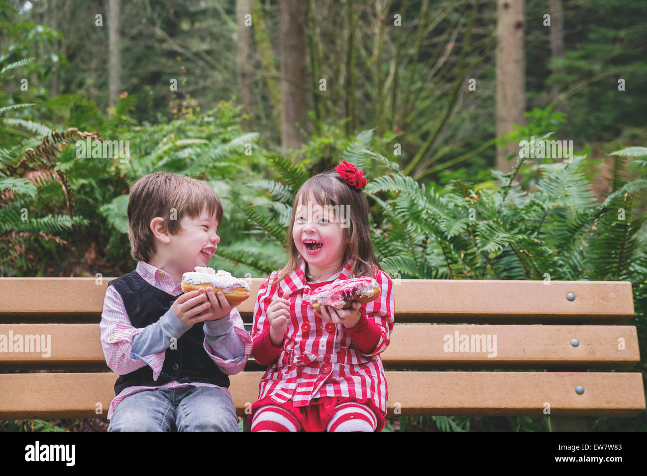 Two children sitting on a bench laughing and eating donuts Stock Photo