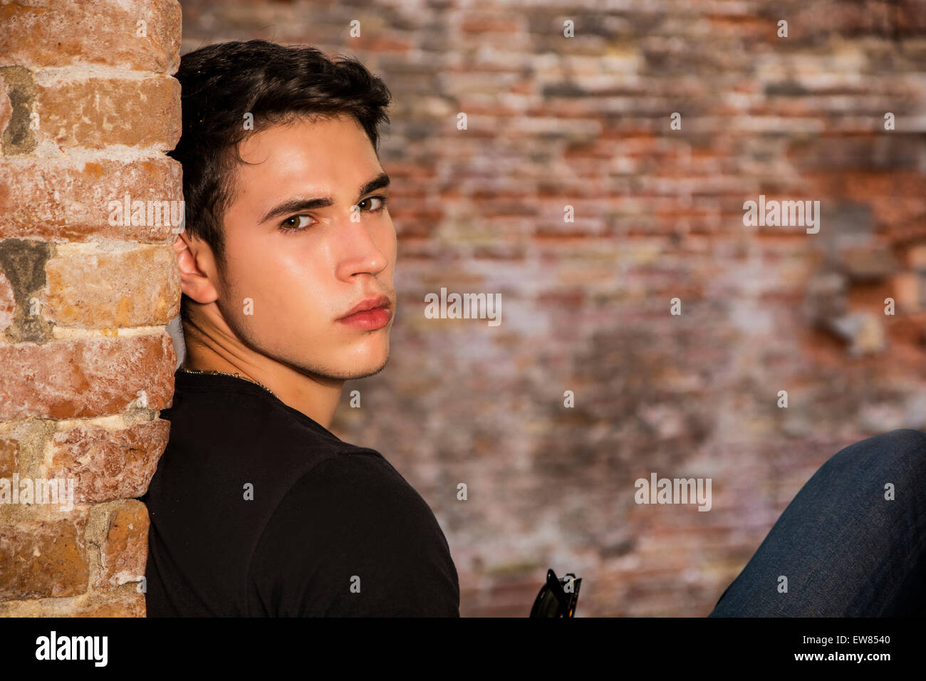 Attractive young man sitting against brick wall, looking at camera Stock Photo