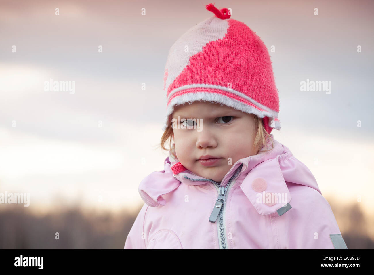 Cute Caucasian blond baby girl in pink hat angry frowns, outdoor closeup portrait Stock Photo