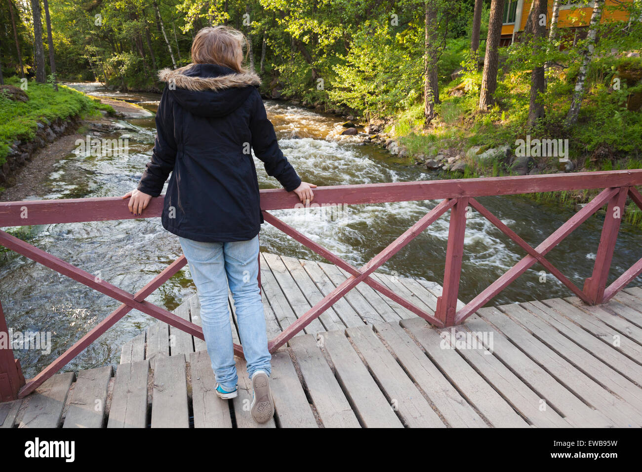 Beautiful blond Caucasian teenage girl stands on wooden bridge across small fast river in Finland, back view Stock Photo