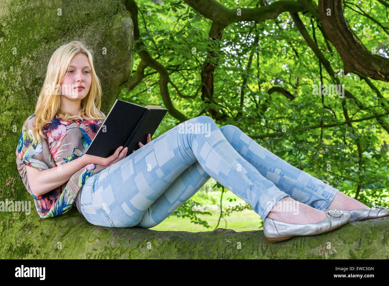 Blonde caucasian teenage girl lying on green tree reading book Stock Photo