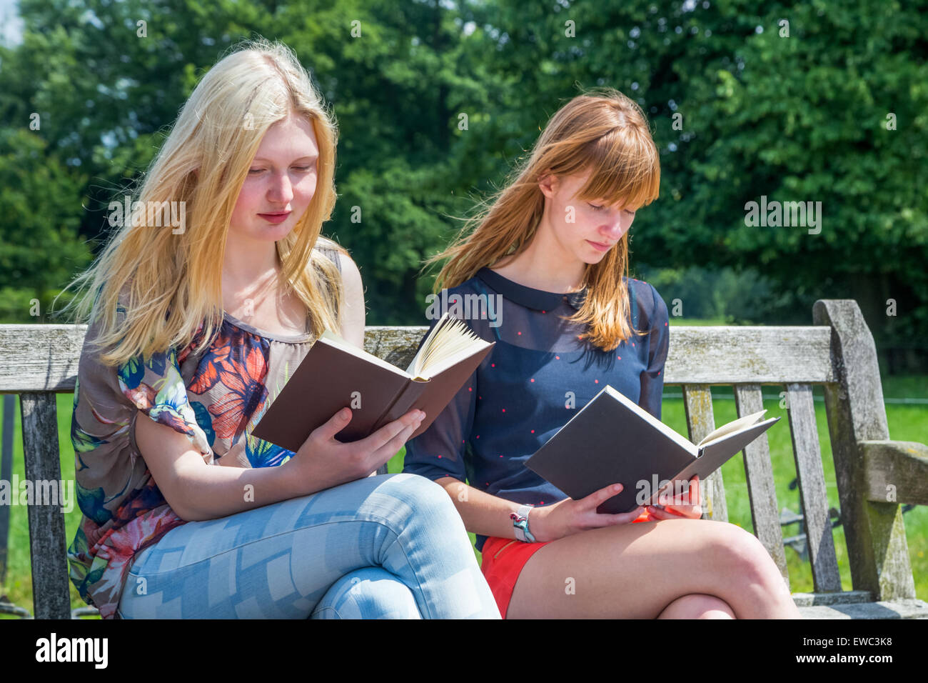 Two caucasian teenage girls reading books on bench in green nature Stock Photo