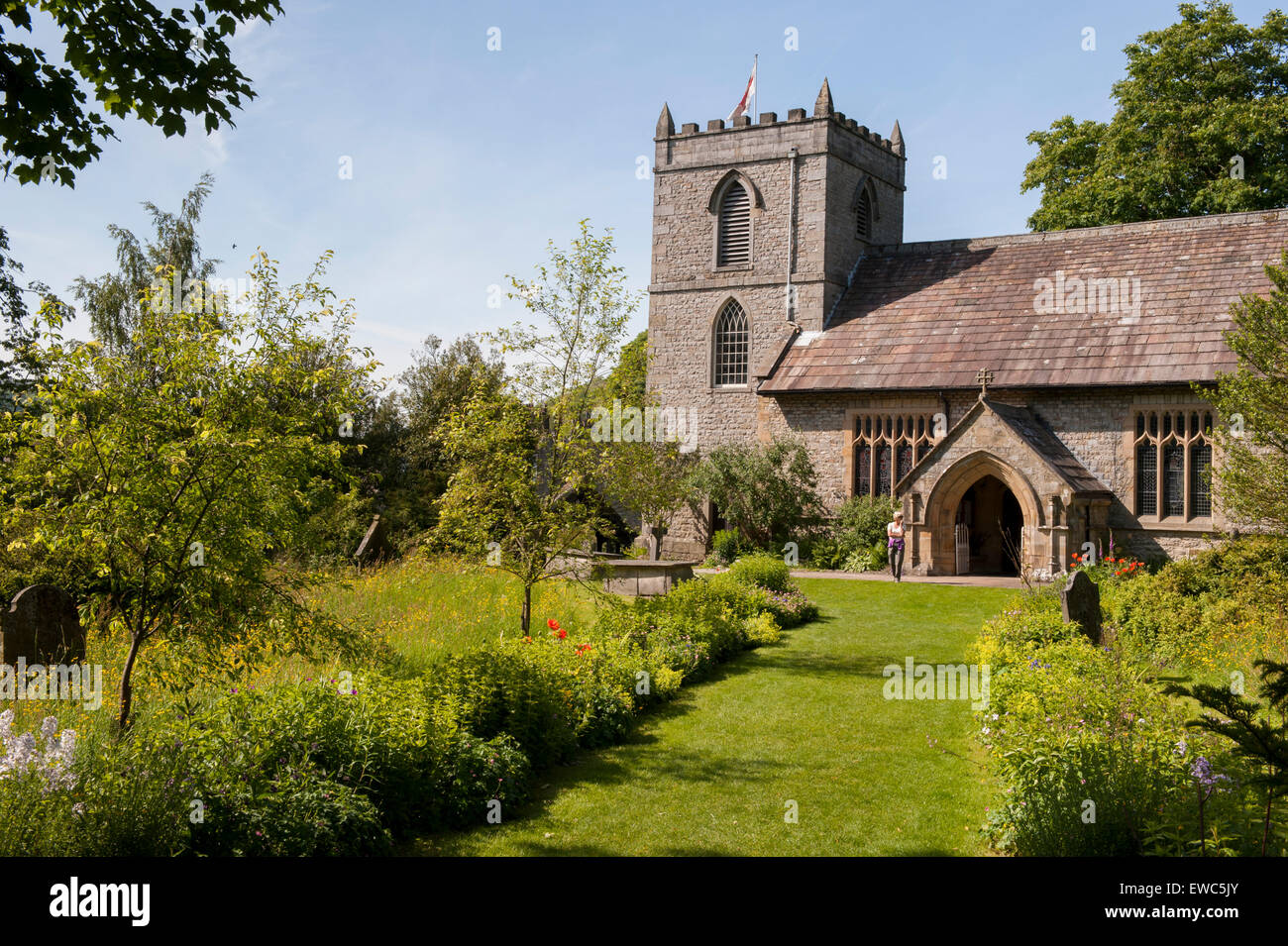 St. Mary's church, Kettlewell, Yorkshire Dales, England, UK. 1 lady standing by porch, beautiful garden with meadow flowers, summer sun & blue sky. Stock Photo