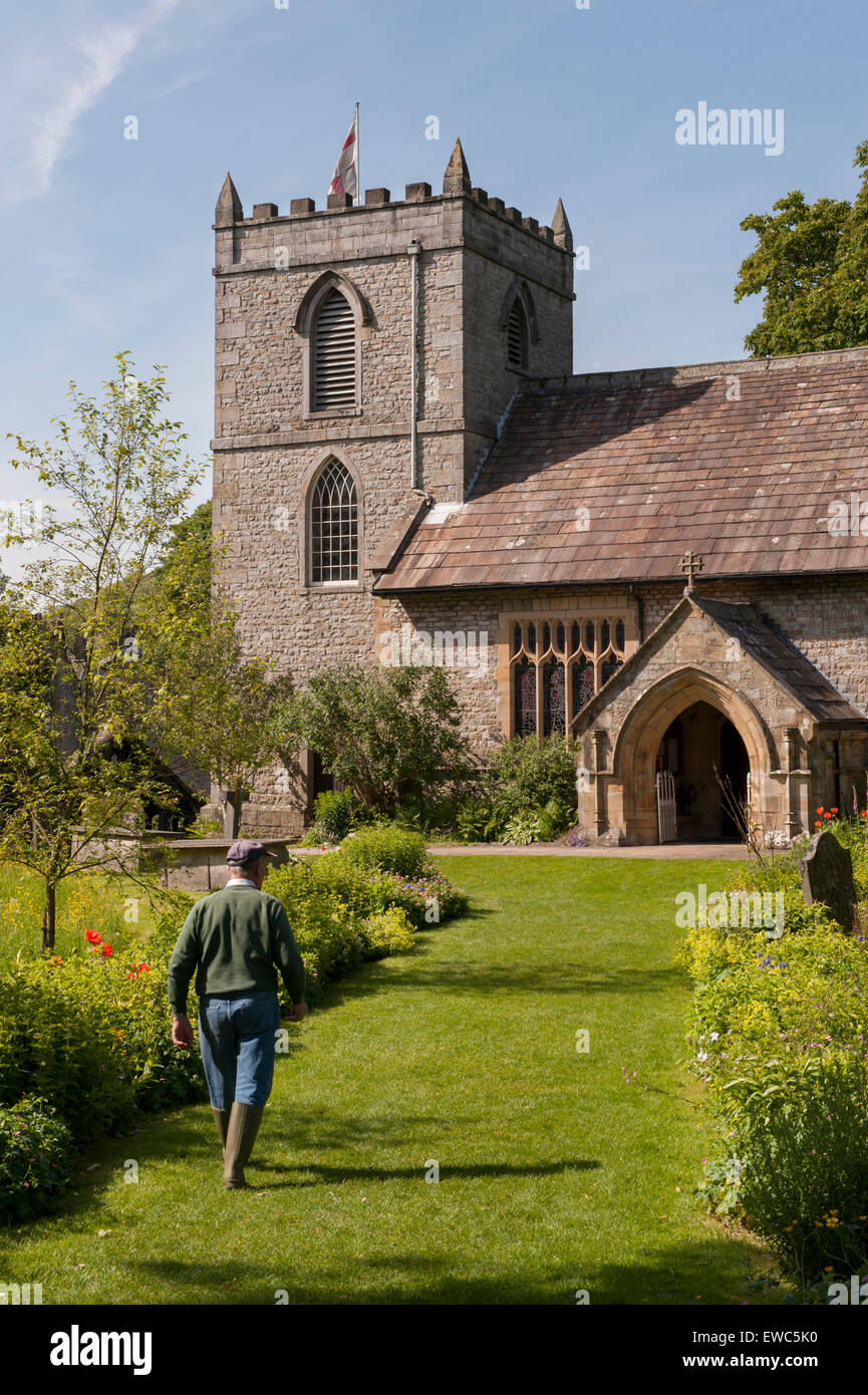 St. Mary's church, Kettlewell, Yorkshire Dales, England, UK. View of gardener on path in beautiful garden with meadow flowers, summer sun & blue sky. Stock Photo