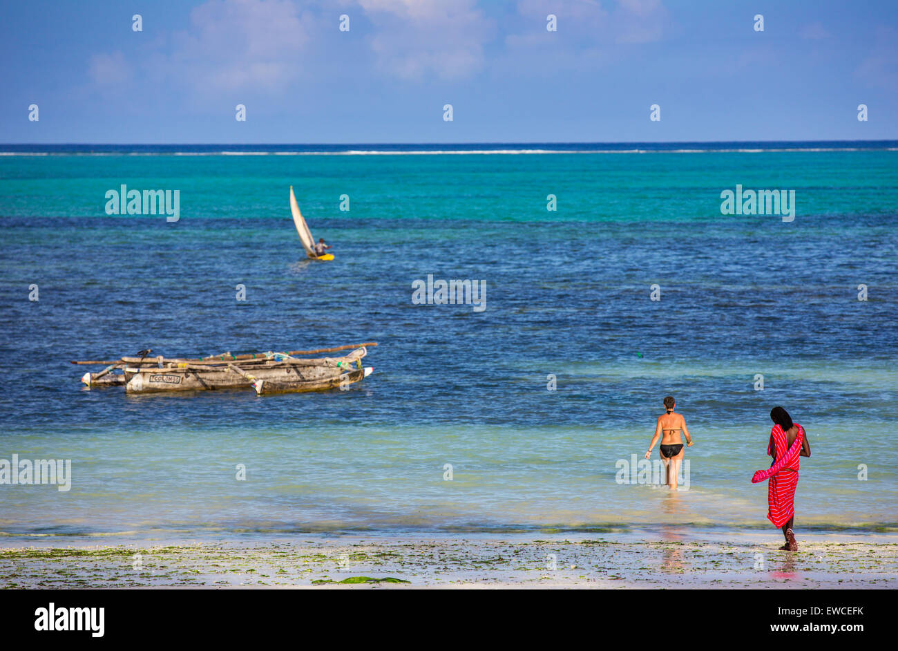 A masai man watches a tourist as she walks into the water in Zanzibar, Tanzania. Stock Photo
