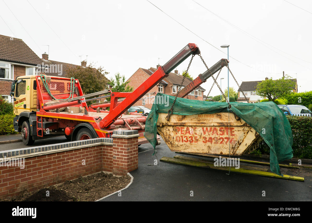 A full +covered waste skip being collected by lorry to a constricted suburban driveway Derby England Stock Photo