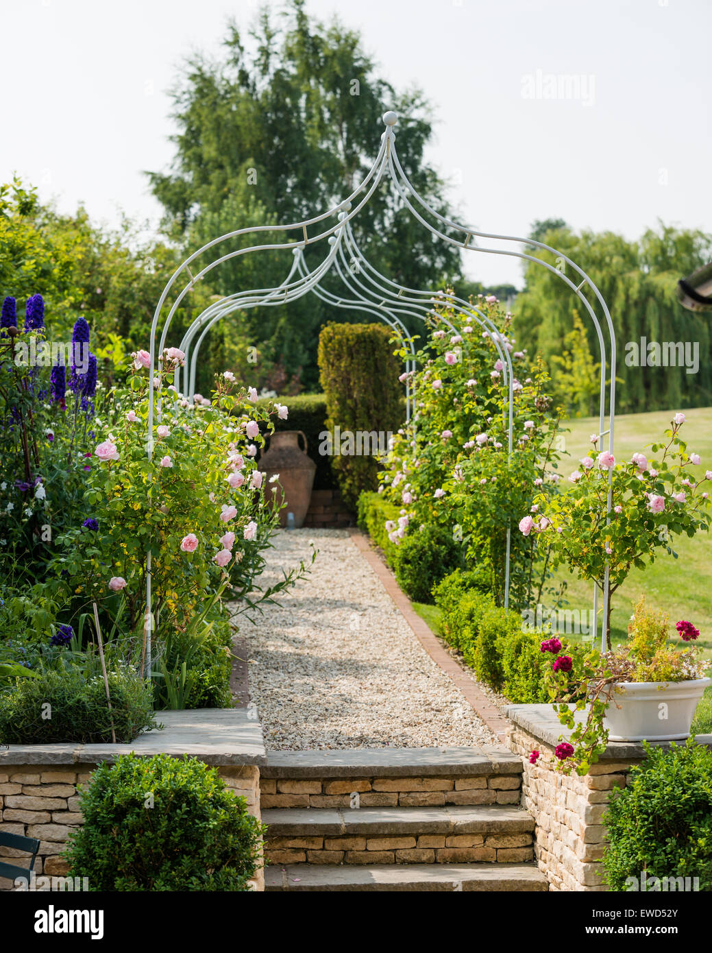 Gravel pathway with wrought iron pergola and flowering rose bushes in garden of Cotswold famhouse Stock Photo