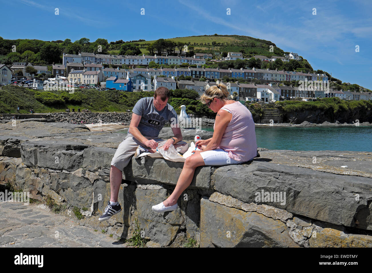 A couple sit on the quay eating fish and chips in hot sunny weather by the beach at New Quay