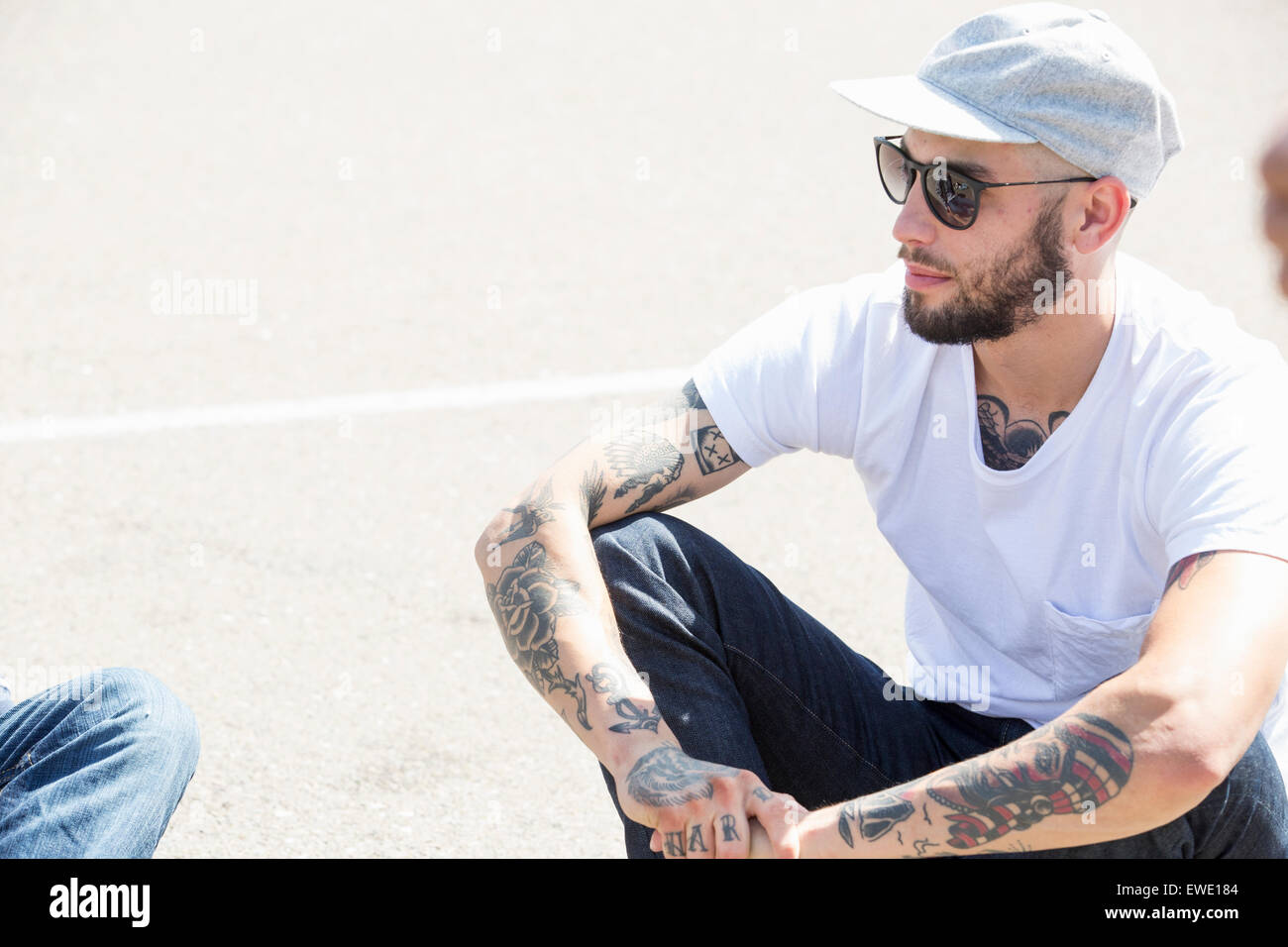 Young man with tattoos, wearing a cap and sunglasses, sitting on the ground Stock Photo