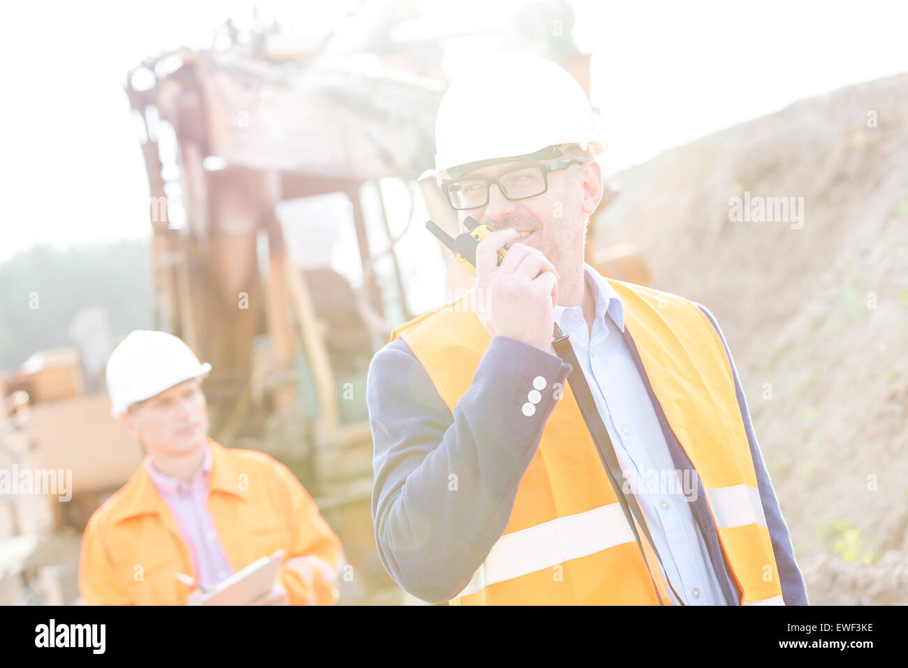 Happy engineer using walkie-talkie on construction site with colleague in background Stock Photo