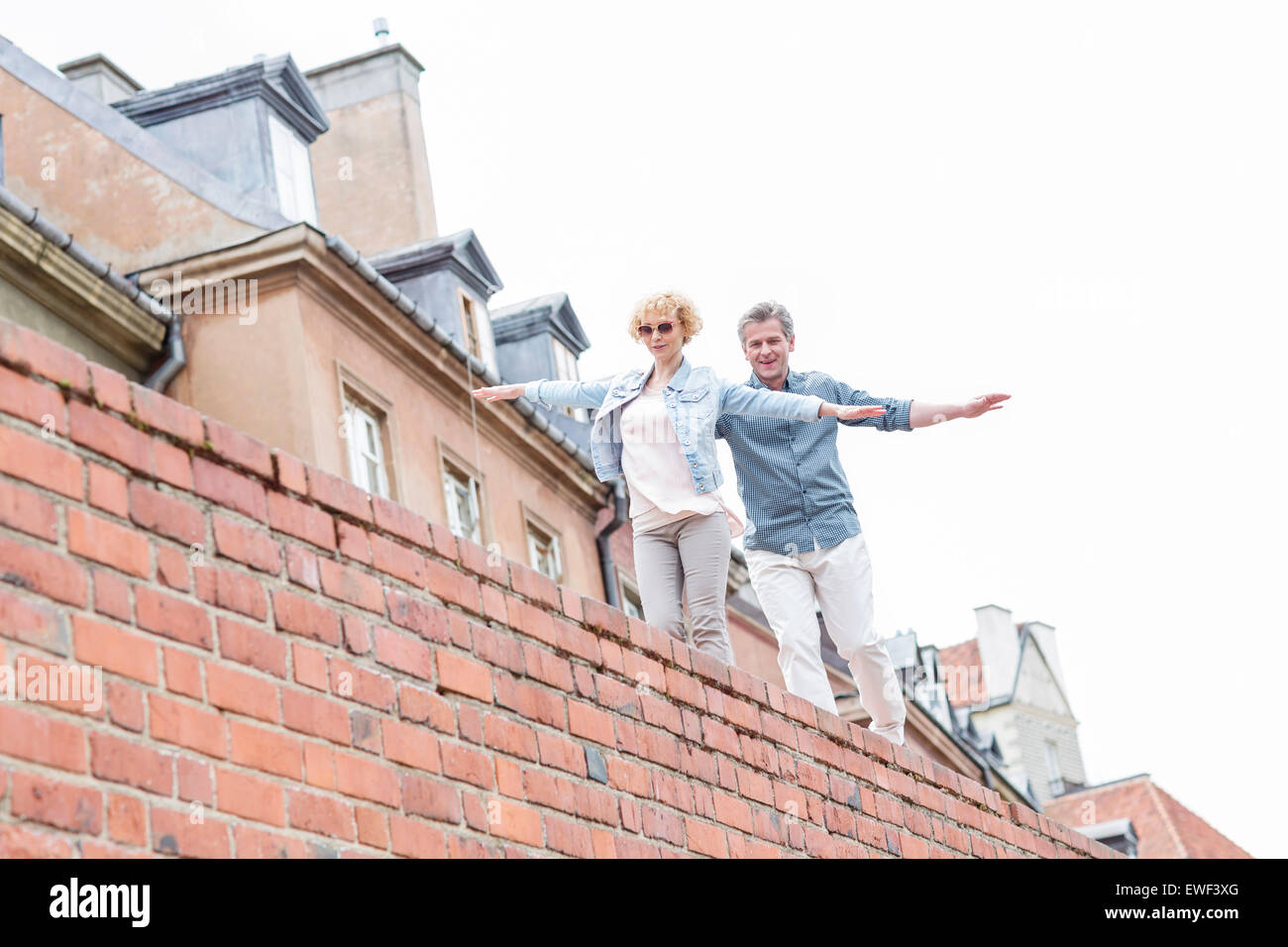 Low angle view of middle-aged couple with arms outstretched walking on brick wall against clear sky Stock Photo