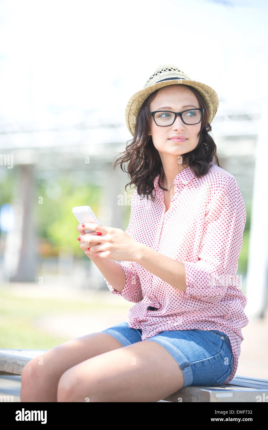 Woman looking away while holding mobile phone on bench outdoors Stock Photo